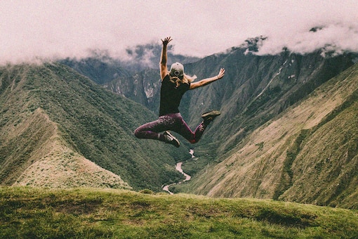 Frau springt in die Luft, Berge, Wolken, Almwiese, Fluss fließt durch ein Tal