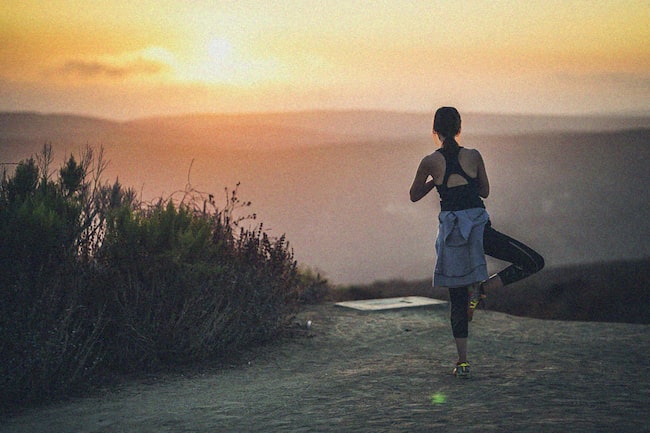 Frau steht auf einem Bein und dehnt sich beim Joggen auf einem Berg. Sie schaut in Richtung eines Sonnenuntergangs und die Stimmung wirk sehr harmonisch.