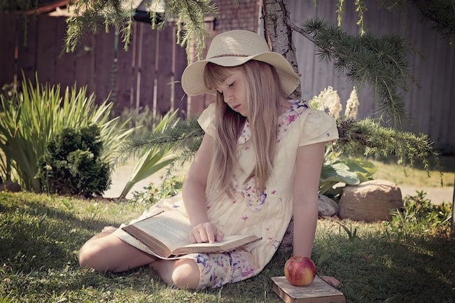 Mädchen mit Sonnenhut und Kleid liest ein Buch im Garten in der Wiese sitzend. Daneben liegt ein Apfel.