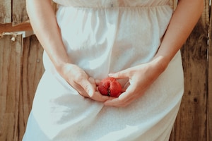 Detailaufnahme von einer Frau im weißen Kleid, die eine Erdbeere vor ihrem Schambereich in den Händen hält